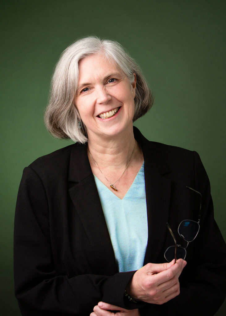 Studio headshot of a smiling, grey haired woman in business attire, holding a pair of glasses.