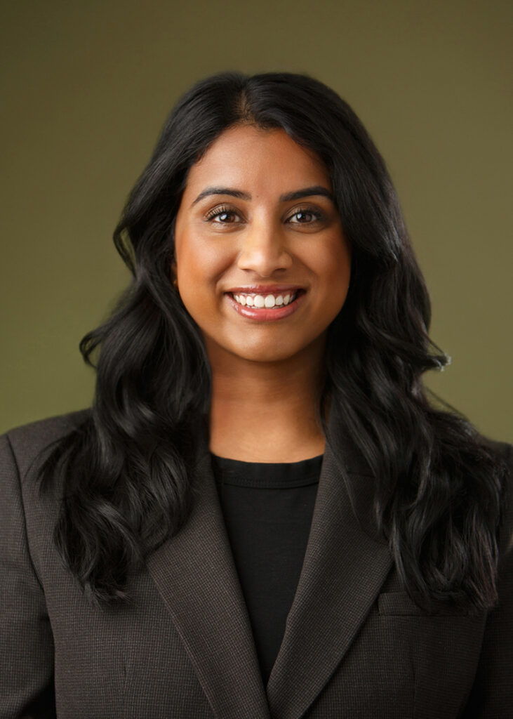 Studio headshot of a smiling, young woman in business attire and long black wavy hair.